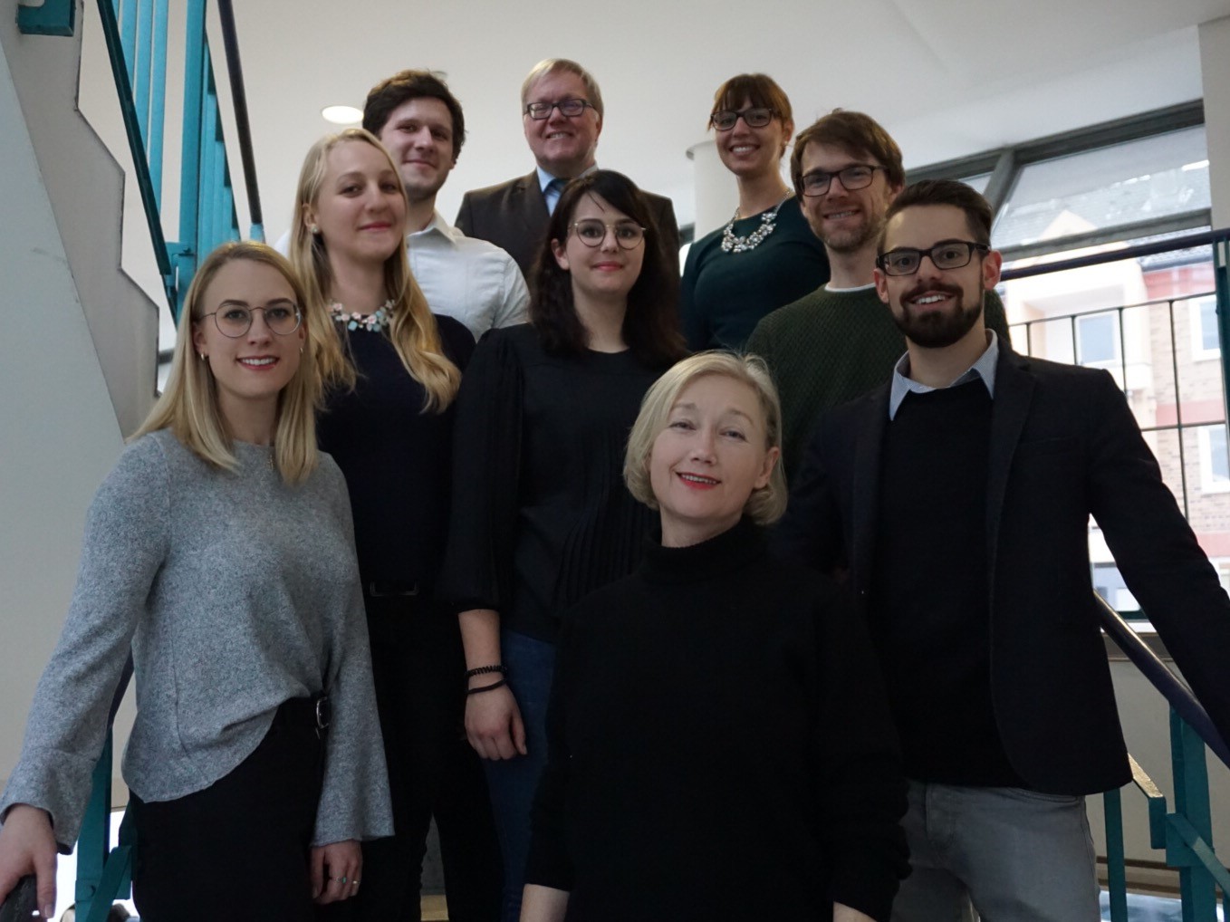 Auf dem Bild ist das Lehrstuhlteam zu sehen. Sie stehen auf der Treppe im Juridicum zum ersten Stock, in drei Reihen a 3 Personen. In der ersten Reihe steht, von links nach rechts: Yvonne Baumgärtner, Ingrid Mümmler und [...]. In der zweiten Reihe steht: [...], Alexandra Lörinczy und Thomas Herbein. In der dritten Reihe steht: [...], Prof. Dr. Max Emanuel Geis und [...].